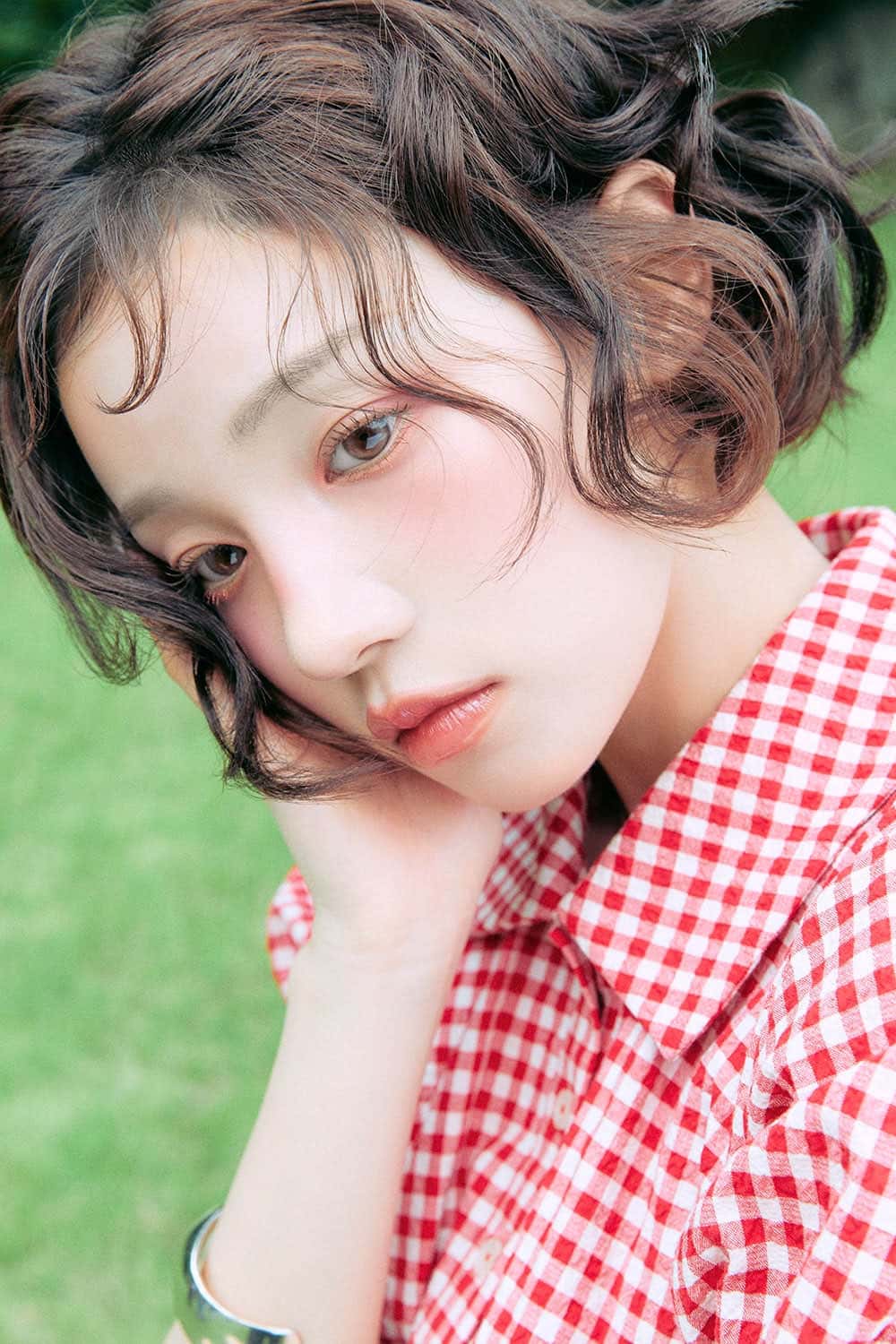 Retrato de prensa de YUQI con el pelo corto y ondulado, camisa a cuadros rojos y blancos y la mano apoyada en la mejilla, sobre un fondo de pasto. Foto: Cube Entertainment