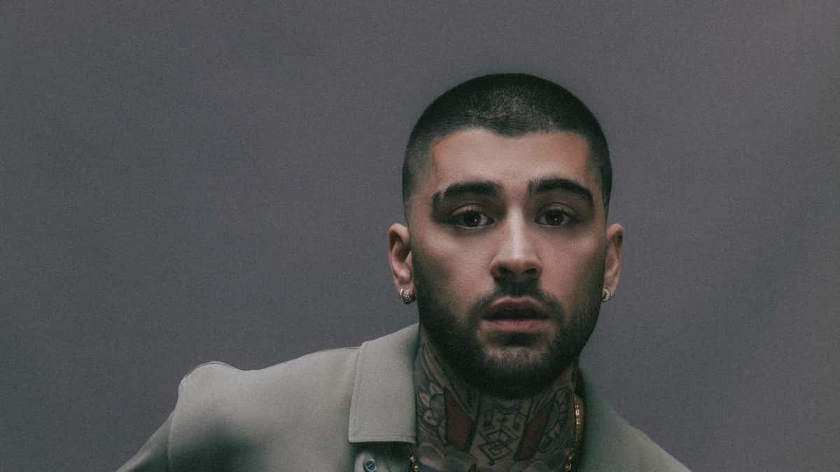 Retrato de prensa de ZAYN con el pelo rapado, barba, aros y tatuajes en el cuello, con camisa verde grisácea frente a una pared gris