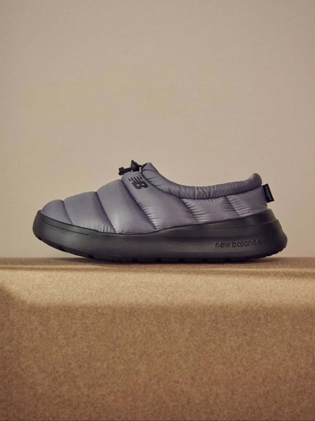 New Balance Sleeping Moc gris, acolchada con costuras horizontales y suela negra, apoyada sobre una alfombra