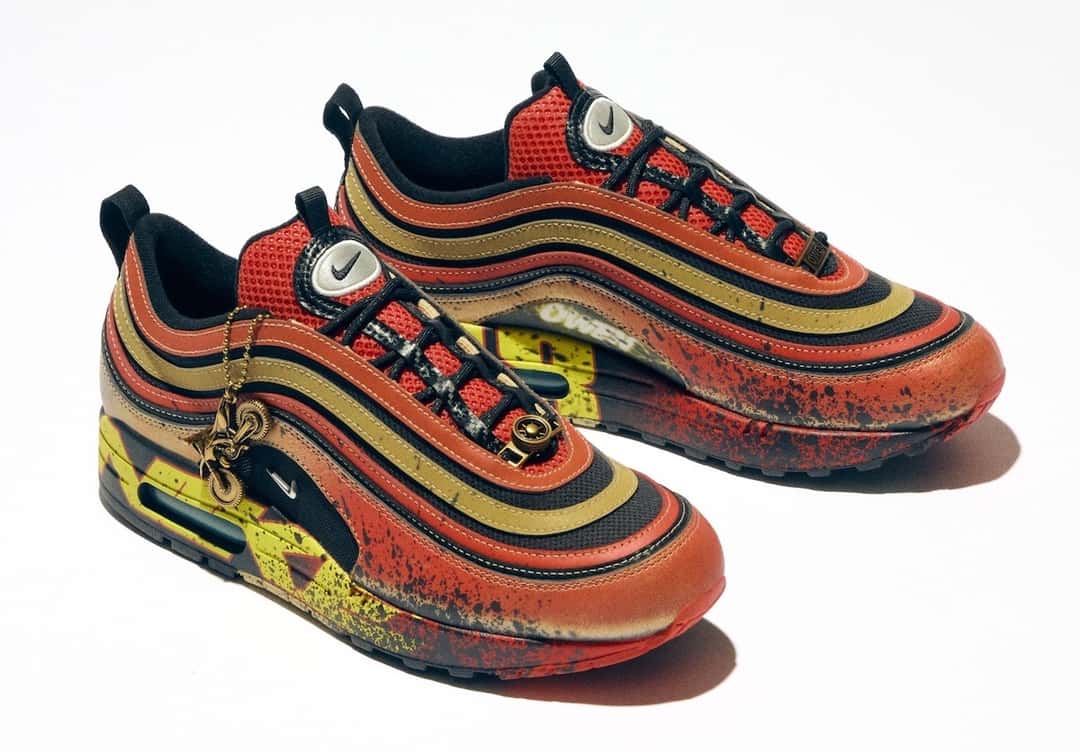 Nike Air Max 1/97 de la colección Doernbecher Freestyle 22 sobre fondo blanco, con las líneas onduladas del Air Max 97 en rojo, dorado y negro, salpicaduras oscuras, una zona amarilla en el talón y un colgante dorado en los cordones