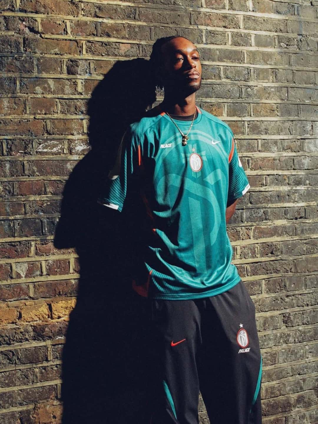Persona apoyada contra una pared de ladrillos con la camiseta verde azulado de Palace x Nike Manor Place FC y el pantalón deportivo negro