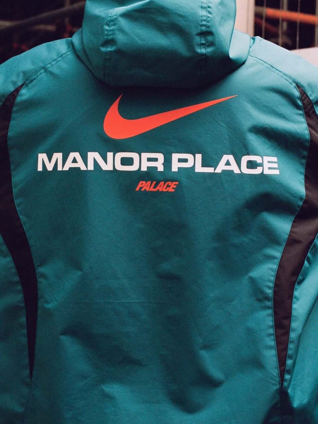 Espalda de una campera verde azulado de Palace x Nike con el Swoosh naranja y la leyenda Manor Place en letras blancas