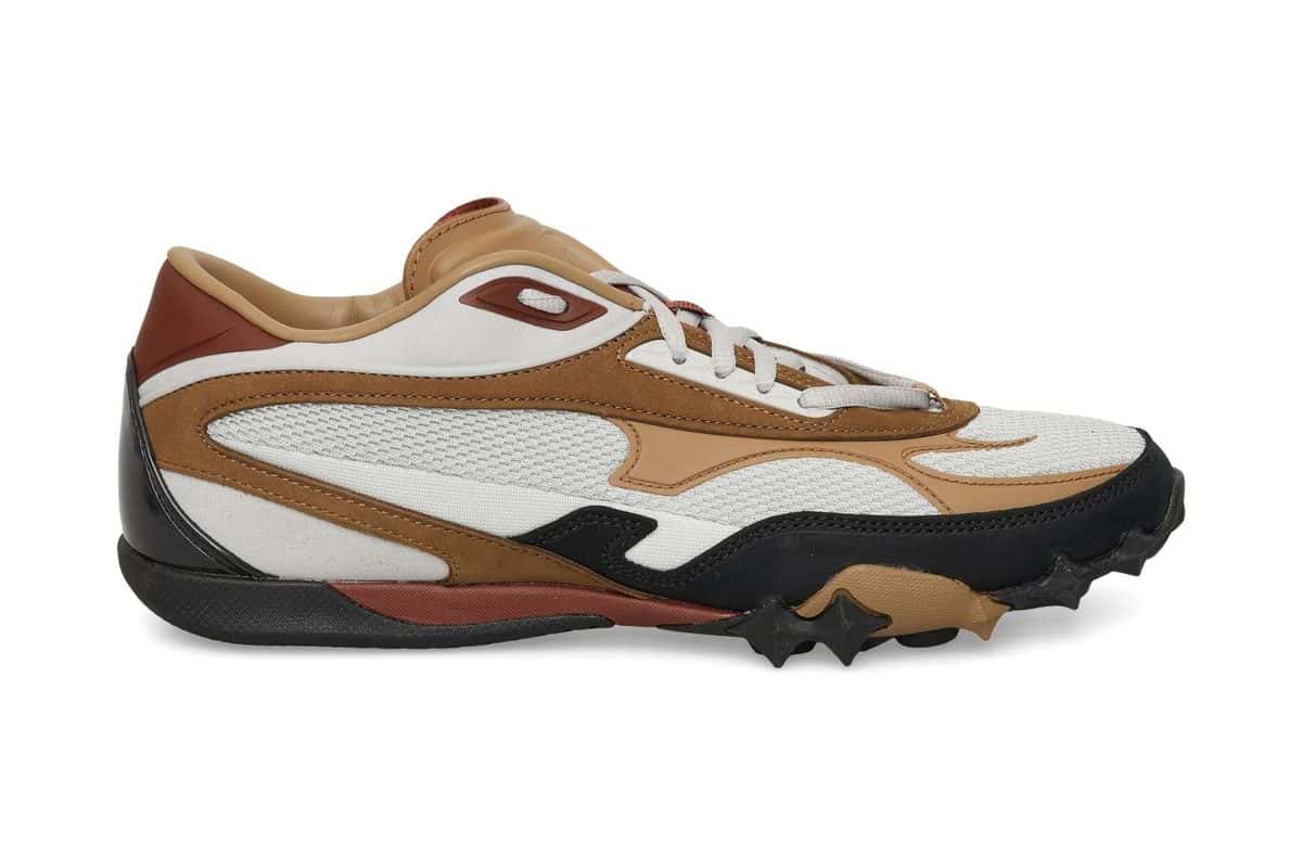 Reebok Spike Racer gris, marrón y negra de perfil sobre fondo blanco, con malla gris clara, piezas de cuero marrón y la suela negra con tacos en la punta