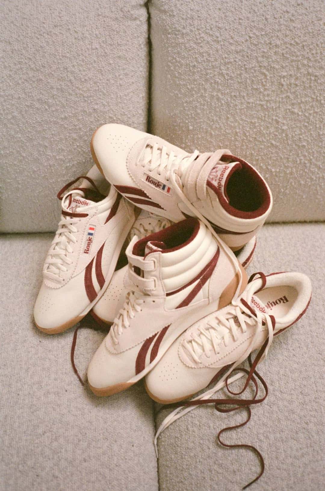 Pares de Reebok Freestyle Hi y Lo de Rouje en crema y bordó apoyados sobre un sillón tapizado, con los cordones sueltos