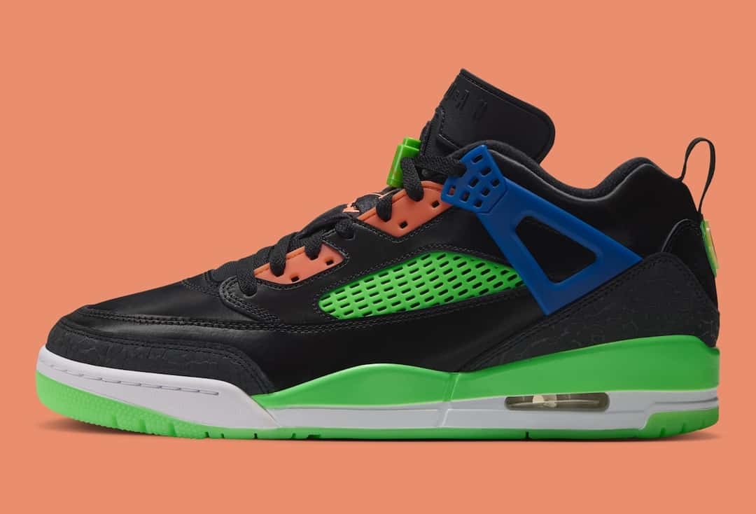 Jordan Spizike Low Jam Central de perfil sobre fondo naranja, con upper de cuero negro, red lateral verde, panel de cordones naranja, ala de cordones azul, entresuela verde y blanca con cámara de aire visible y suela verde