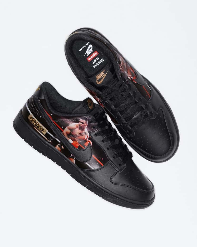 Par del Supreme x Martine Rose x Nike Dunk Low Tyson visto desde arriba sobre fondo blanco, con un zapato mostrando la plantilla negra con los logos de Nike, Supreme y Martine Rose, la etiqueta dorada de Nike en la lengüeta y el otro de costado con la foto de Tyson y las letras doradas