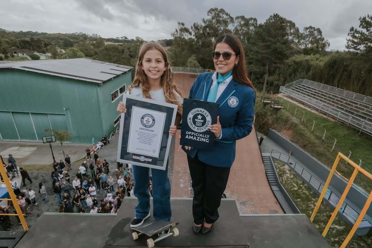 Laura Voigt sostiene su certificado del Guinness junto a una representante de Guinness World Records, parada con su tabla en lo alto de la megarrampa, con el público abajo