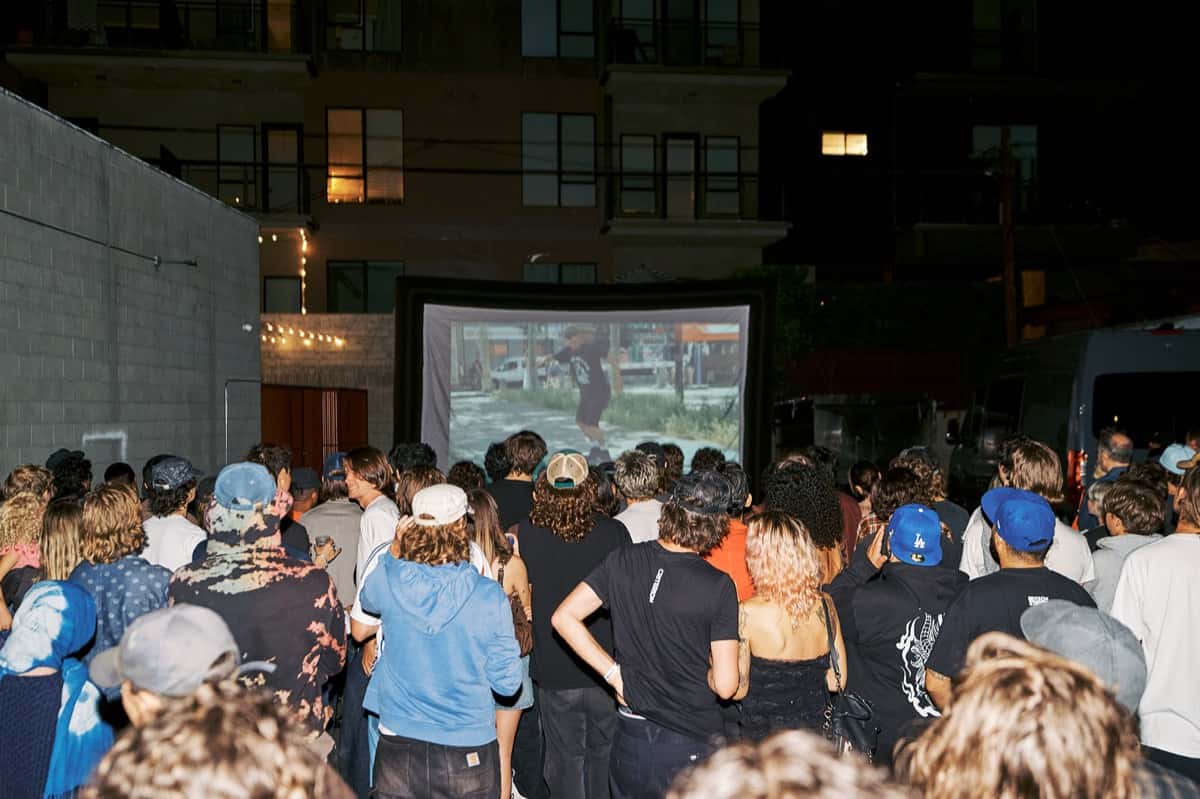 Público de espaldas mira en una pantalla al aire libre el estreno de «Eighty-Nine» en el patio del OBEY Art Space de Los Ángeles, de noche