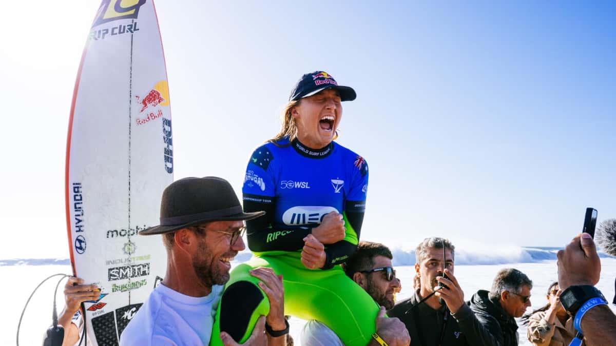 Molly Picklum, con la lycra azul de la WSL, una gorra azul y el traje verde, grita de emoción mientras la llevan en andas por la arena de Supertubos, al lado de su tabla
