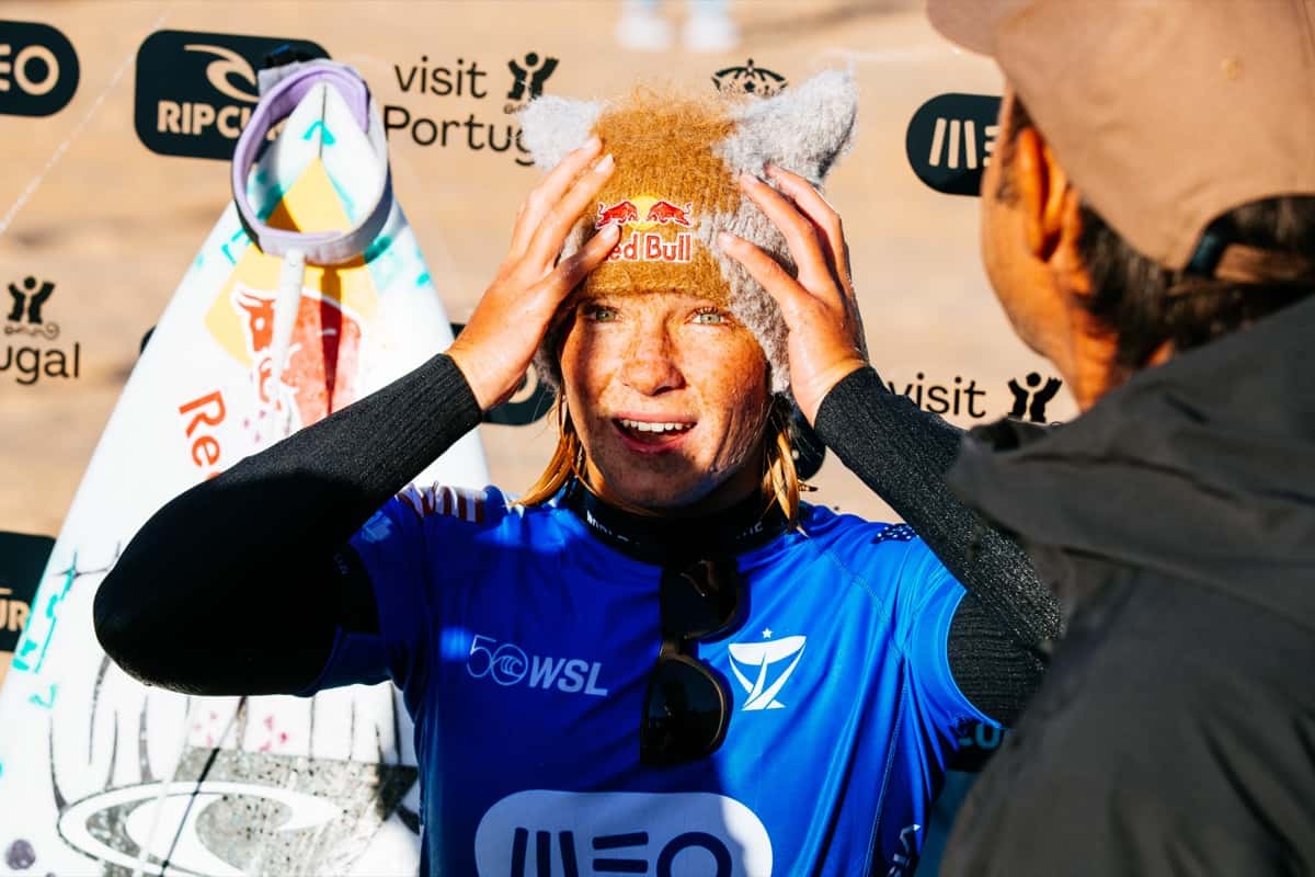 Caitlin Simmers, con la lycra azul de la WSL y un gorro de lana con orejas, se agarra la cabeza con las dos manos y sonríe en la zona de competidores de Supertubos