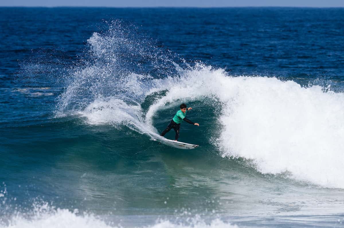 Lucca Mesinas, con lycra verde, tira un giro cerrado en la pared de una ola azul y levanta una cortina de agua, en una fecha de la WSL en Ballito (Sudáfrica) en 2026 (foto de archivo)