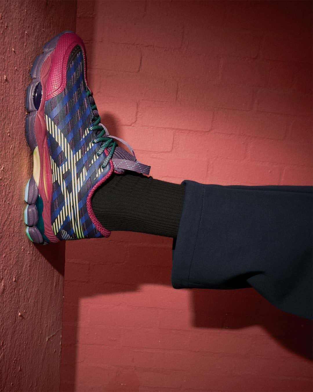 Pierna con media negra y pantalón azul marino apoyada contra una pared de ladrillo pintada de rojo, con la ASICS US8-S GEL-SJ33 Deep Plum/Fresh Grass de perfil en azul y violeta con franjas lima, refuerzos bordó y cordones verdes