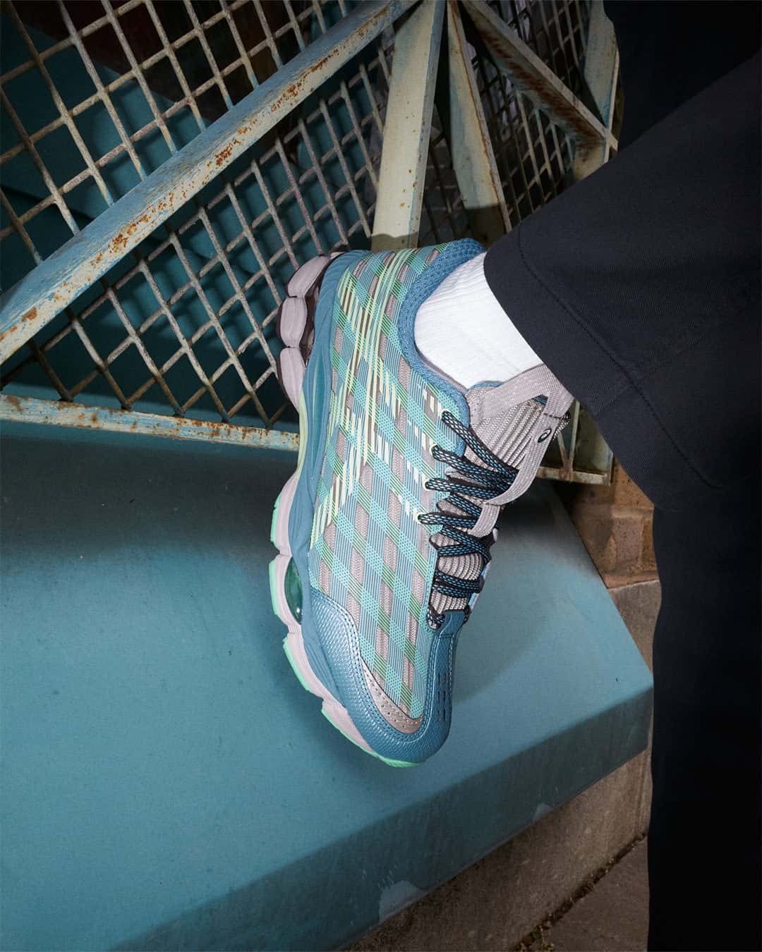 Pie apoyado sobre un escalón turquesa junto a una baranda de reja metálica oxidada, con la ASICS US8-S GEL-SJ33 Moonrock/Tatami Green en azul petróleo, cuadrícula verde agua y lima, cordones oscuros jaspeados, media blanca y pantalón negro