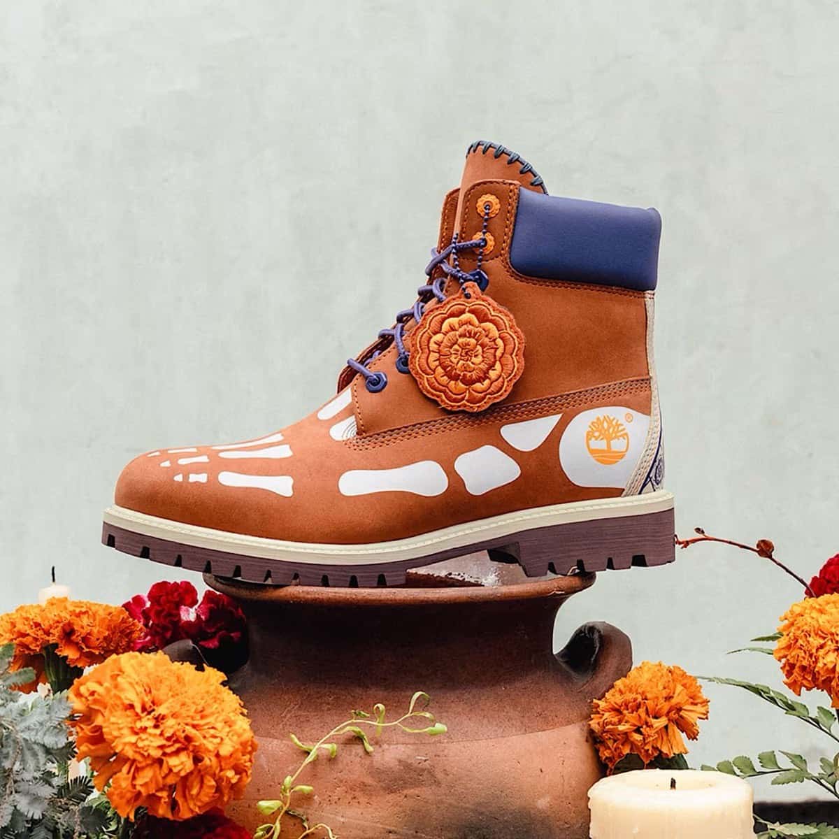 Bota Timberland 6-Inch Día de Muertos en nobuk naranja oscuro con huesos blancos pintados, cuello violeta y un colgante bordado con forma de flor, entre flores de cempasúchil
