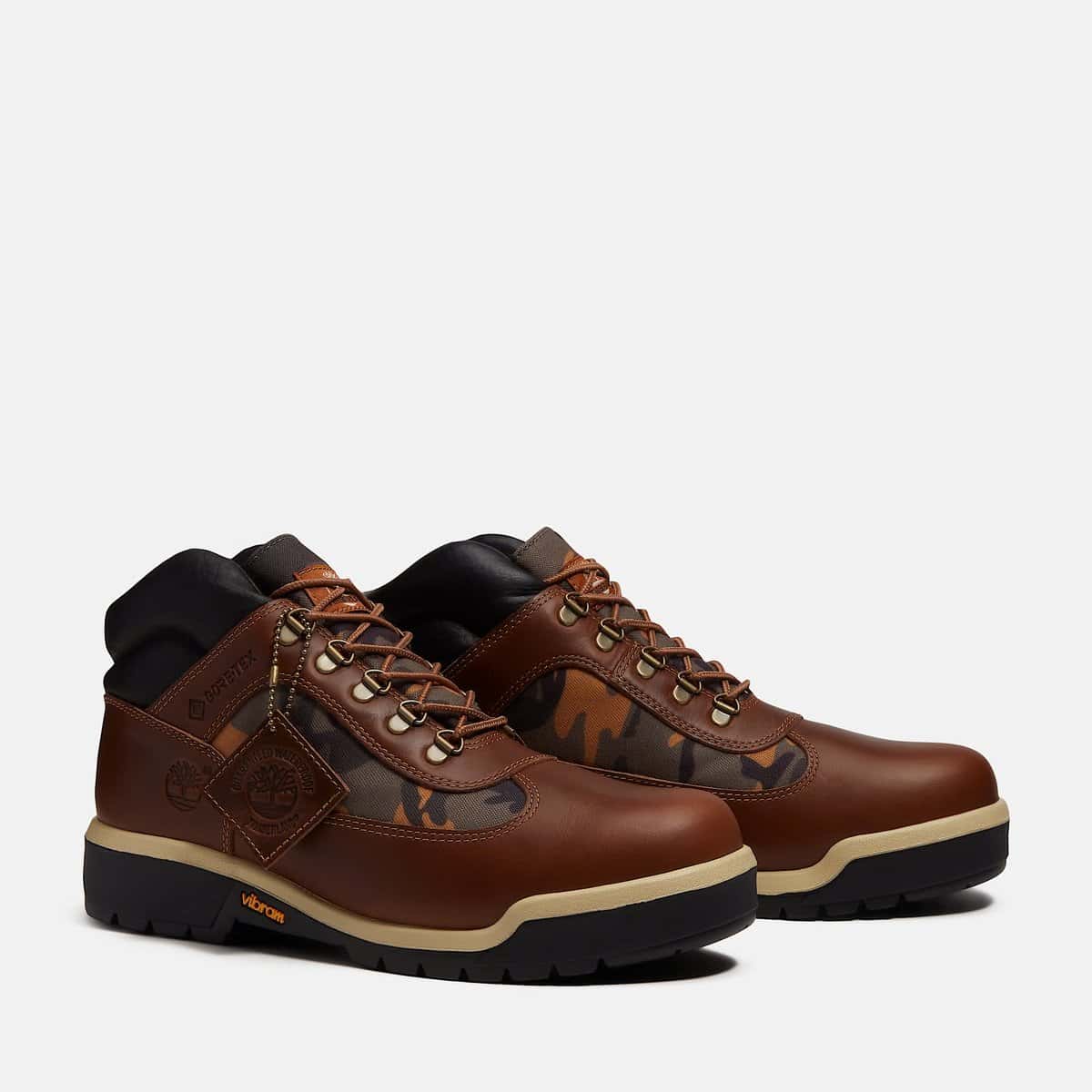 Par de botas Timberland Field Boot con GORE-TEX en cuero marrón, paneles camuflados, cuello acolchado negro, ganchos metálicos y suela Vibram negra con franja crema, sobre fondo gris claro