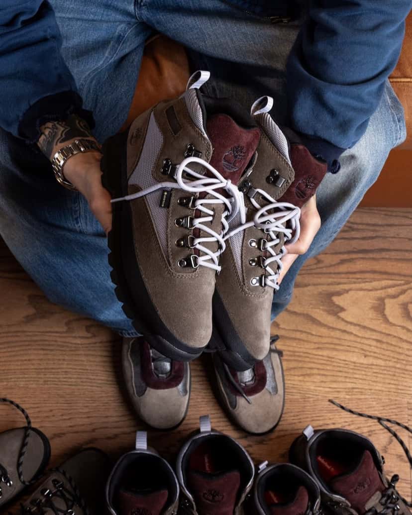 Persona sentada sostiene con las dos manos un par del Victor Kan x Timberland Euro Hiker Gore-Tex en gamuza taupe con malla gris, lengüeta bordó y cordones gris claro, mientras tiene otro par puesto y varias botas iguales en el piso de madera
